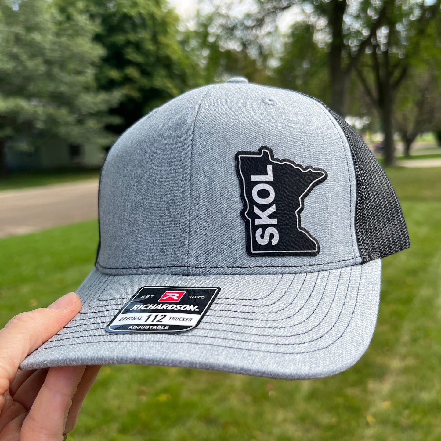 Heather Gray and Black Trucker Hat with an outlined state of Minnesota patch on the left side in black with SKOL engraved vertically on the patch