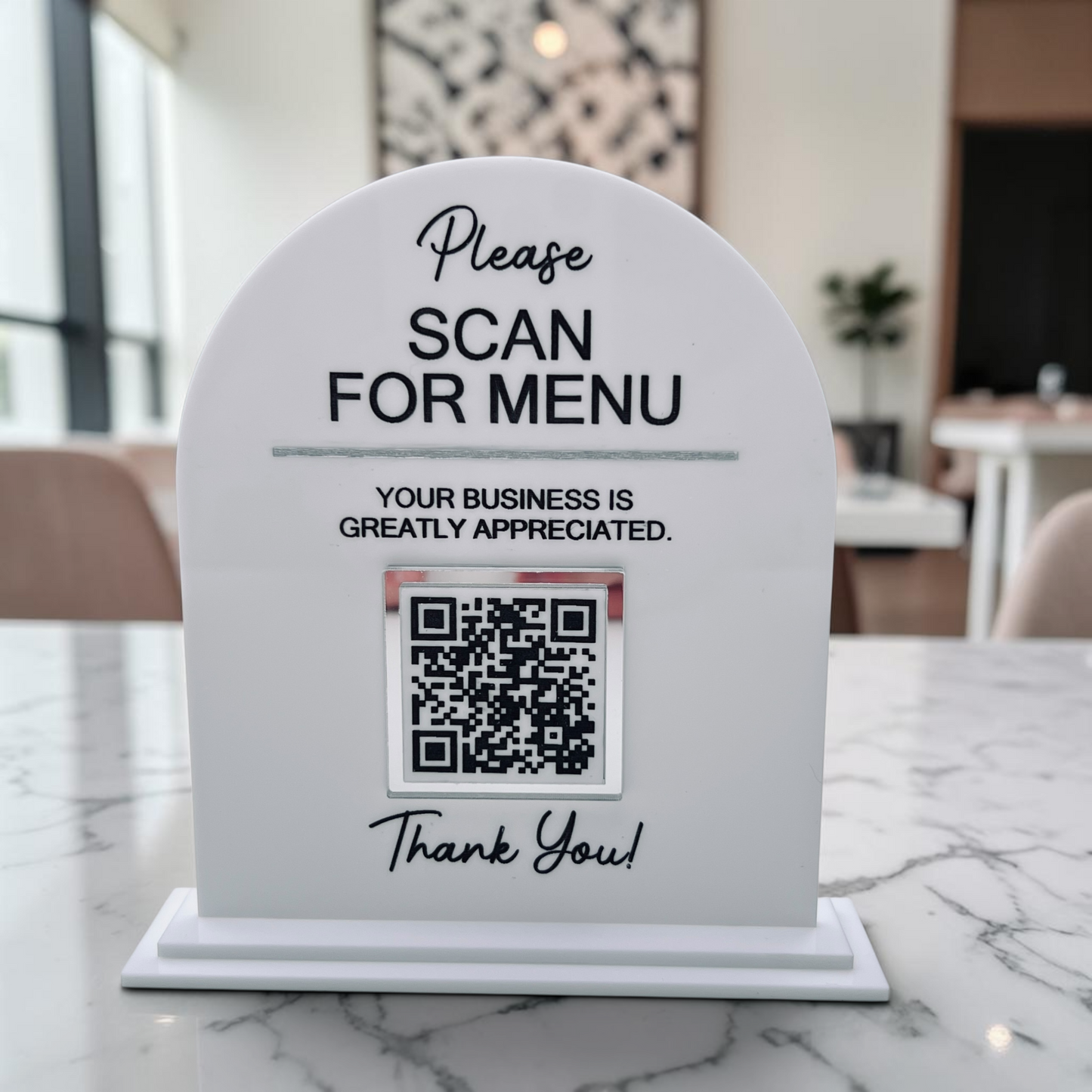 A white arched tabletop sign with a QR code and the text 'Please SCAN FOR MENU' and 'Your business is greatly appreciated. Thank You!' placed on a marble counter in a restaurant setting.