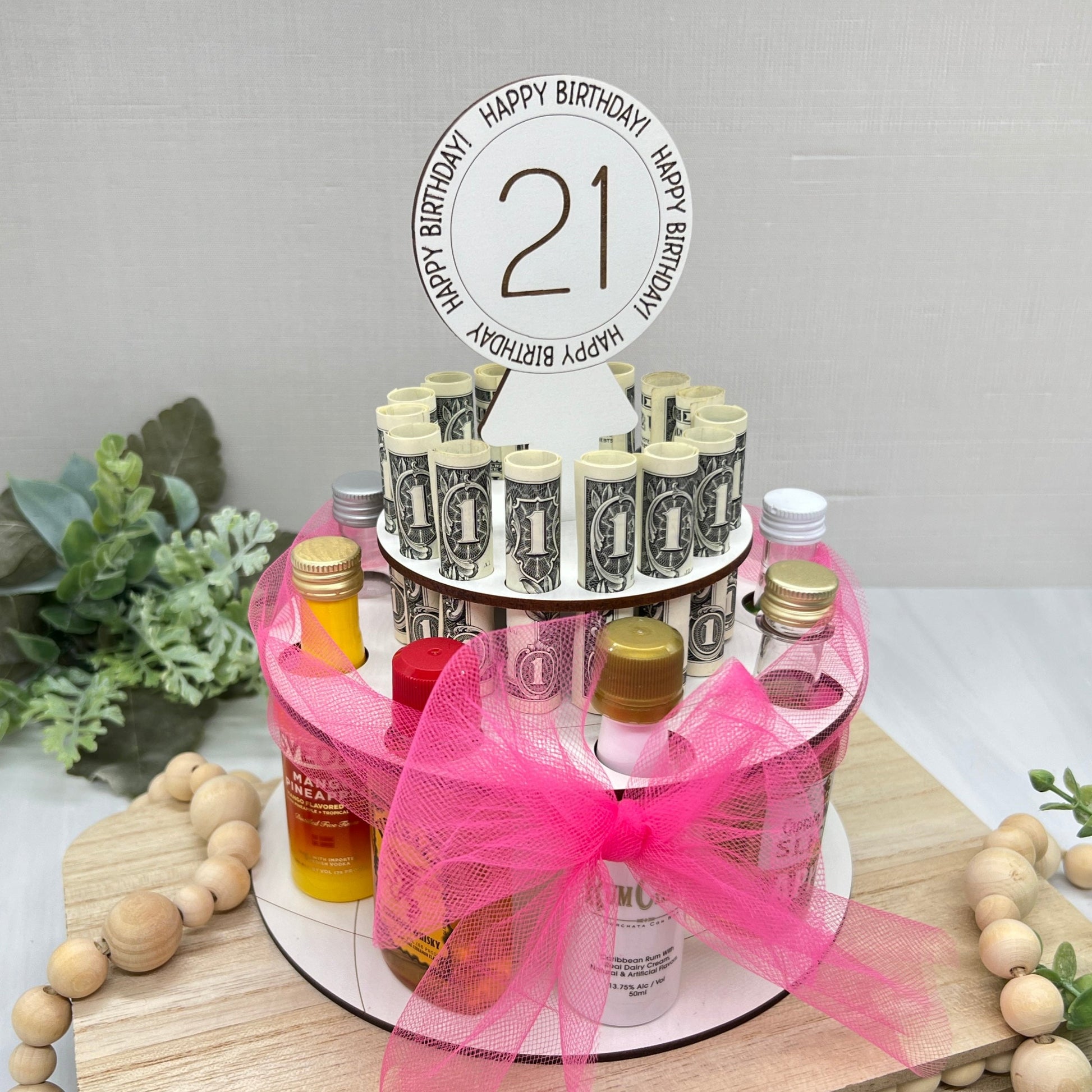 Round 2 layer cake holding 8 mini liquor bottles and rolled up money with a Happy 21st birthday cake topper in white.