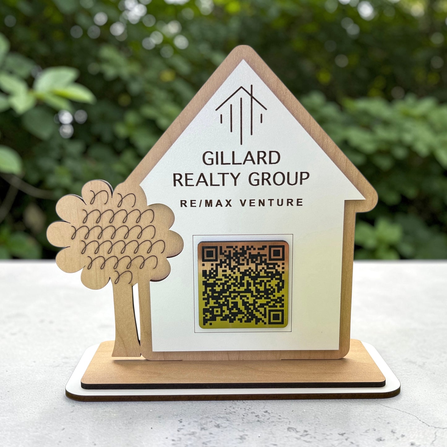 Wooden house-shaped sign with 'Gillard Realty Group' branding and a QR code on a white surface with greenery in the background.