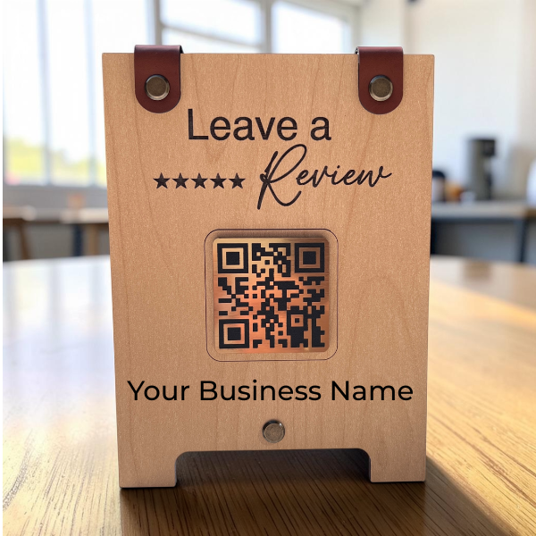A wooden QR code review display standing on a wooden table in a cafe or office setting. The light natural wood front features black printed text reading 'Leave a 5-star Review' and 'Your Business Name', with a central square QR code window and two brown leather strap details at the top.