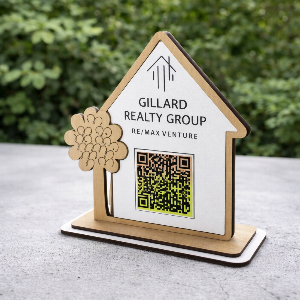 Wooden house-shaped sign with Gillard Realty Group logo and QR code on a surface with greenery in the background