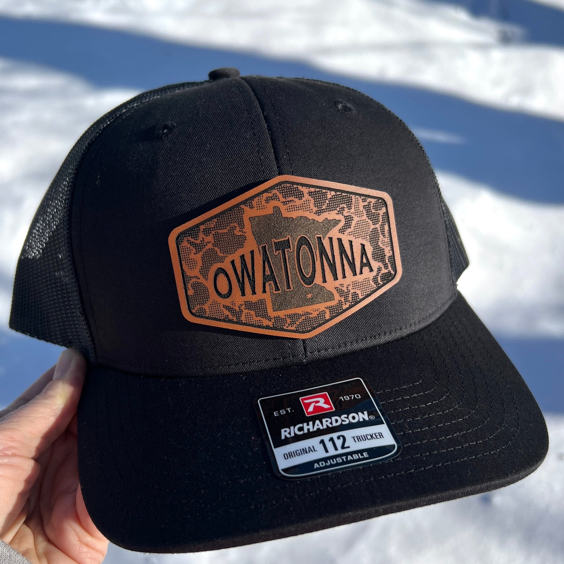 Black Richardson 112 trucker cap with 'Owatonna' leather patch held against a snowy background