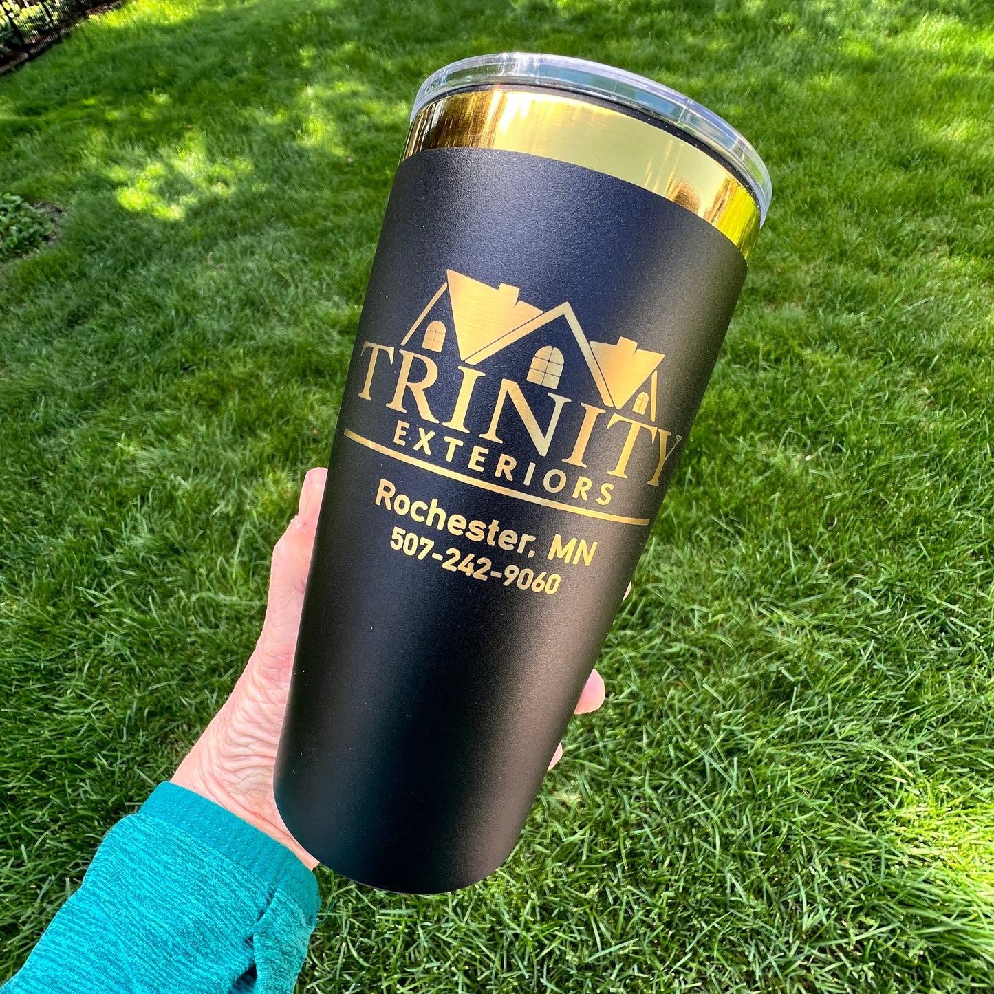 Black tumbler with gold text held in a hand on grass