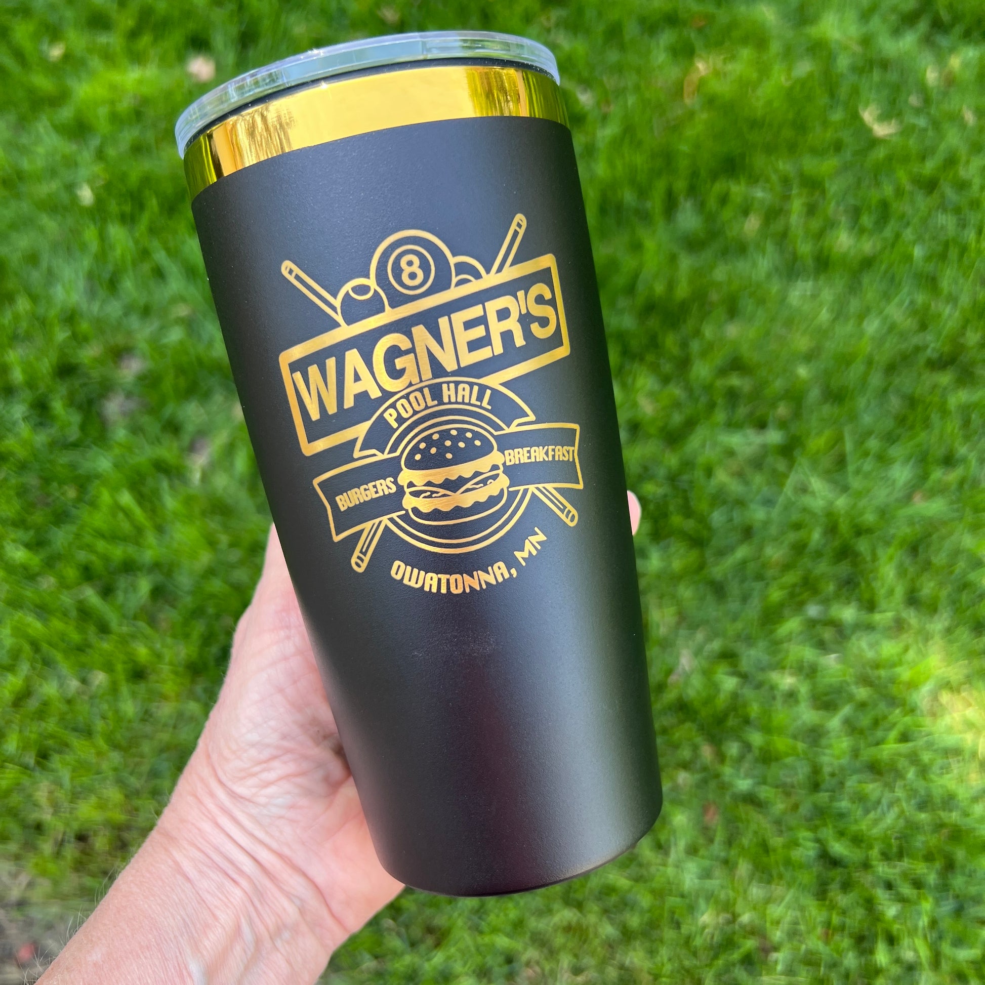 Black tumbler with gold lid featuring 'Wagner's' branding held against a grass background