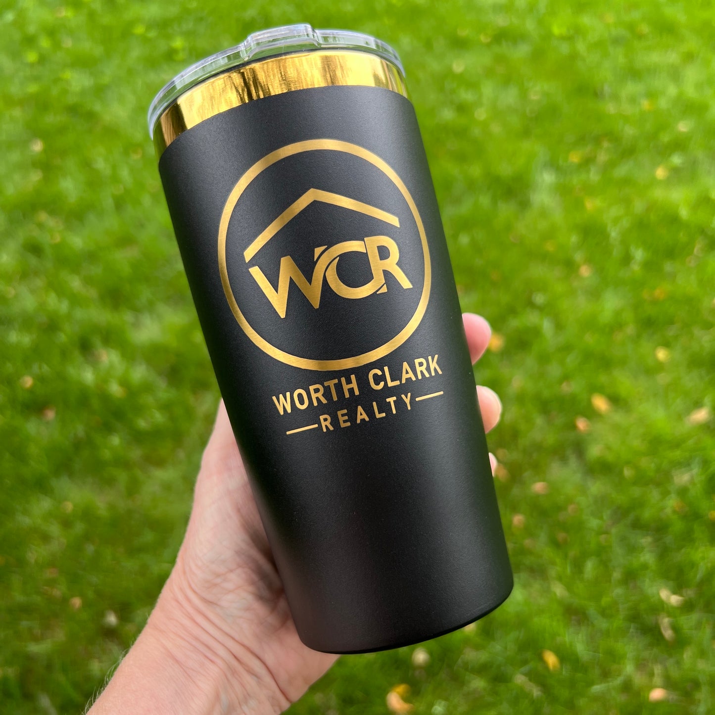 Black tumbler with gold logo and text held in a hand against a green grass background
