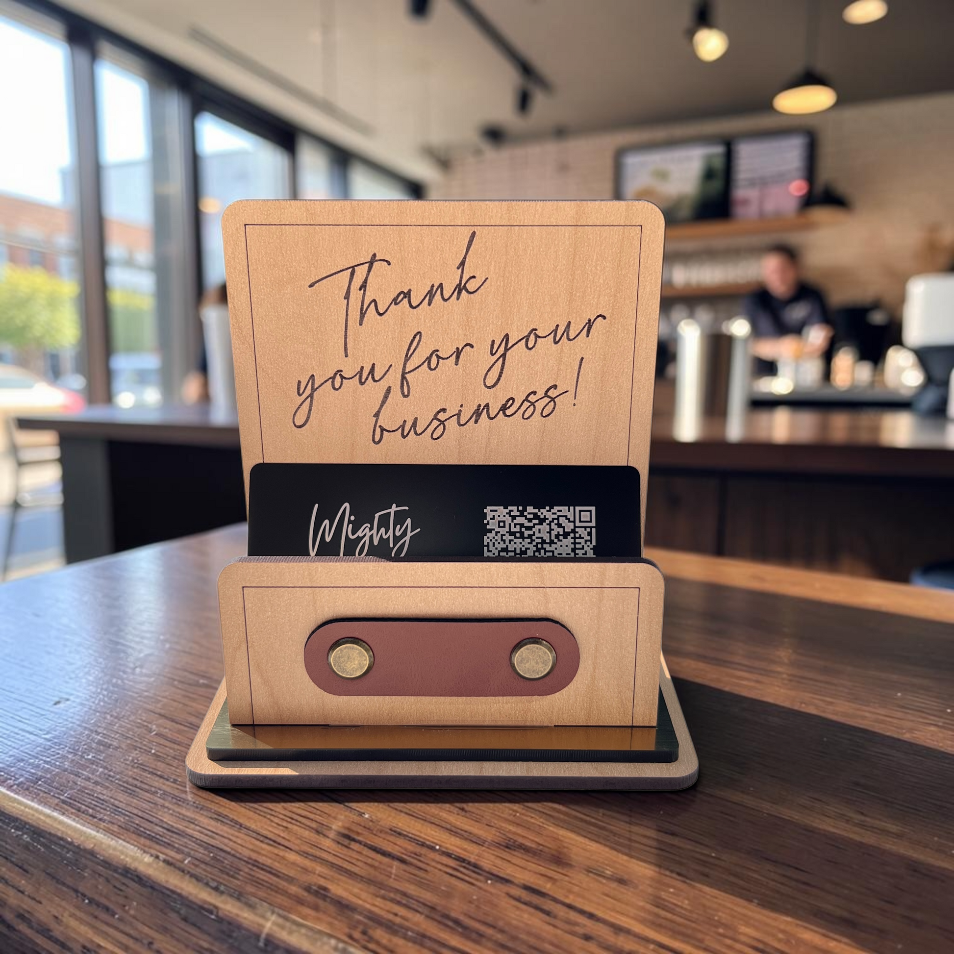A wooden tabletop business card holder with a sign reading 'Thank you for your business!' and a card slot labeled 'Mighty' with a QR code, set on a wooden counter in a cafe with a blurred background.