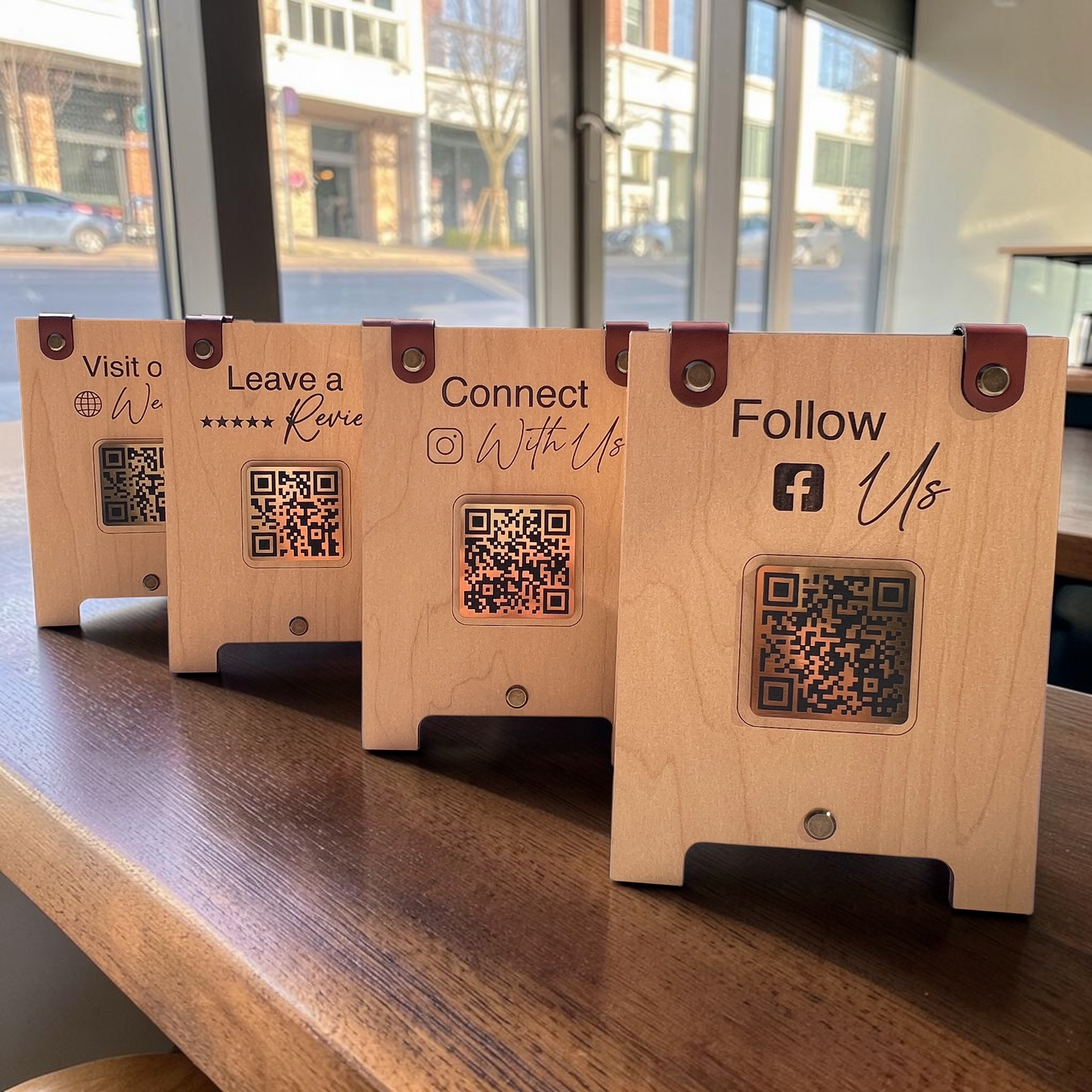 Four wooden QR code display stands arranged in a row on a wooden counter in front of a window. Each stand has a different printed message in black: 'Visit our', 'Leave a Review', 'Connect with Us', and 'Follow Us', with a square QR code cutout in the center and brown leather-like corner straps.