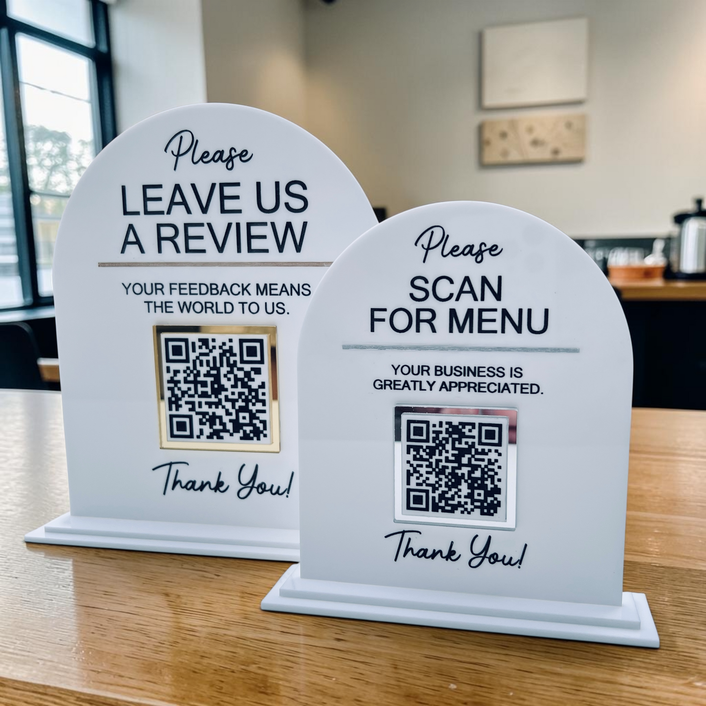 Two white arched plastic review and menu sign stands on a wooden table in a cafe setting. One sign says 'LEAVE US A REVIEW' with a QR code, and the other says 'SCAN FOR MENU' with a QR code.