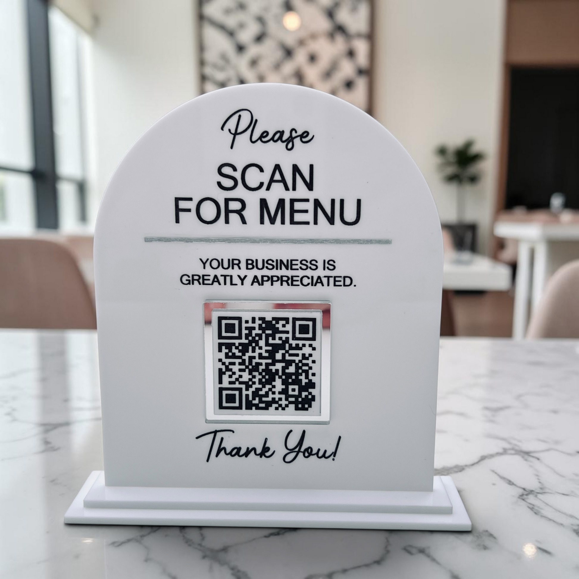 A white arched tabletop sign with a QR code and the text 'Please SCAN FOR MENU' and 'Your business is greatly appreciated. Thank You!' placed on a marble counter in a restaurant setting.