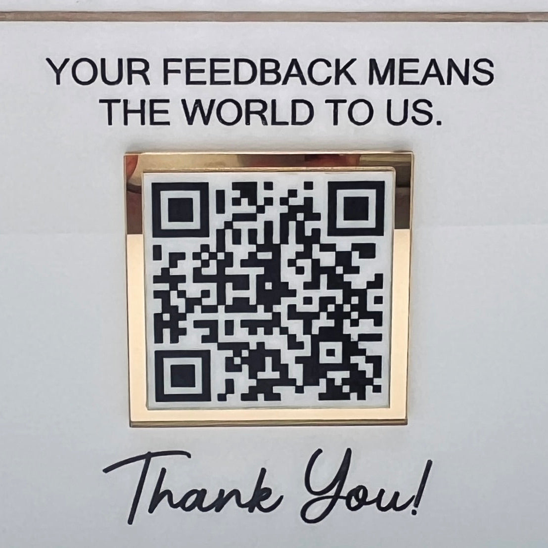 A square acrylic QR code plaque with a gold-toned metallic frame, mounted on a white wall below the text 'YOUR FEEDBACK MEANS THE WORLD TO US.' and above 'Thank You!' in black script.
