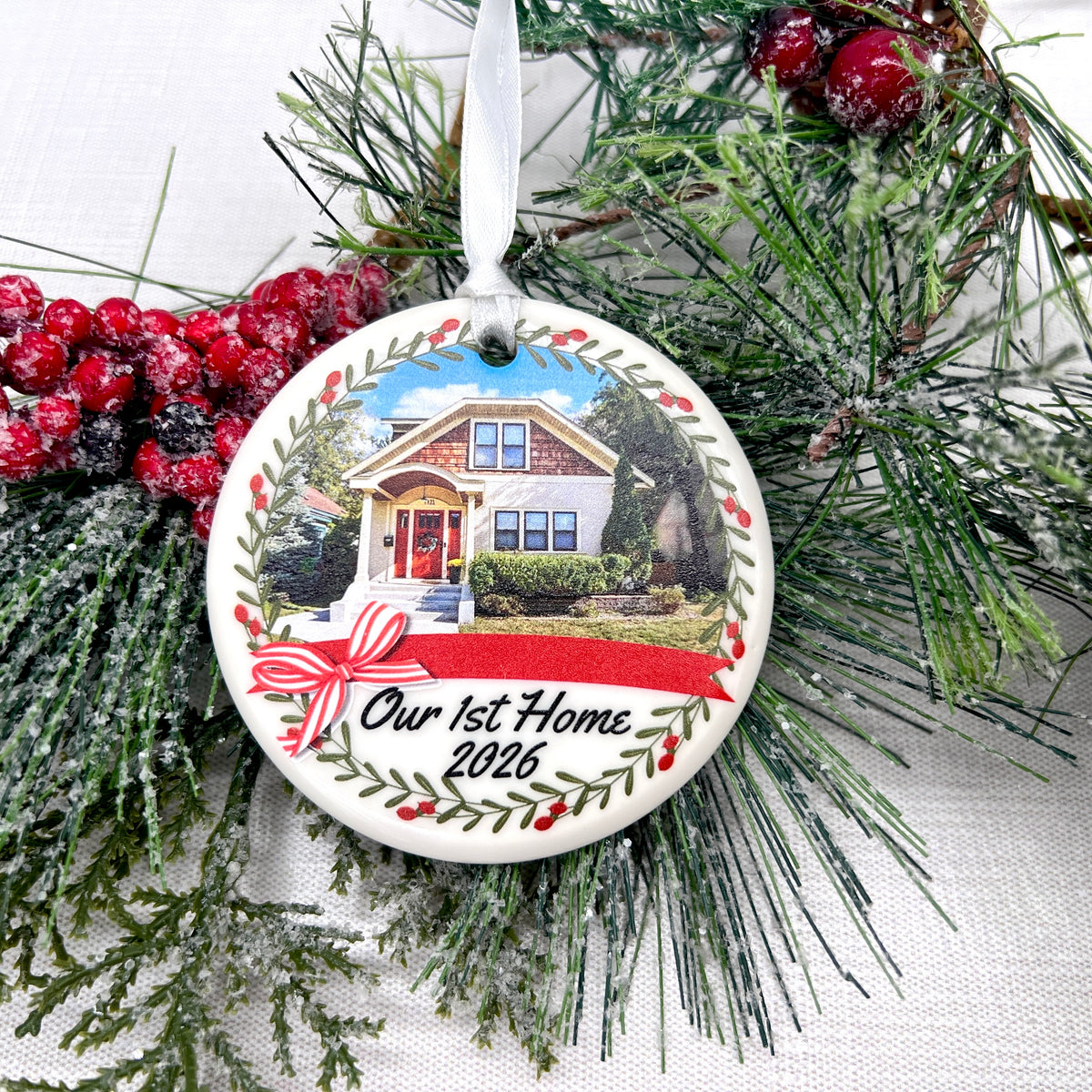 A round ceramic Christmas ornament featuring a printed illustration of a house with the text 'Our 1st Home 2026' and a red ribbon bow, hanging from a white ribbon amidst green pine branches and red berries.