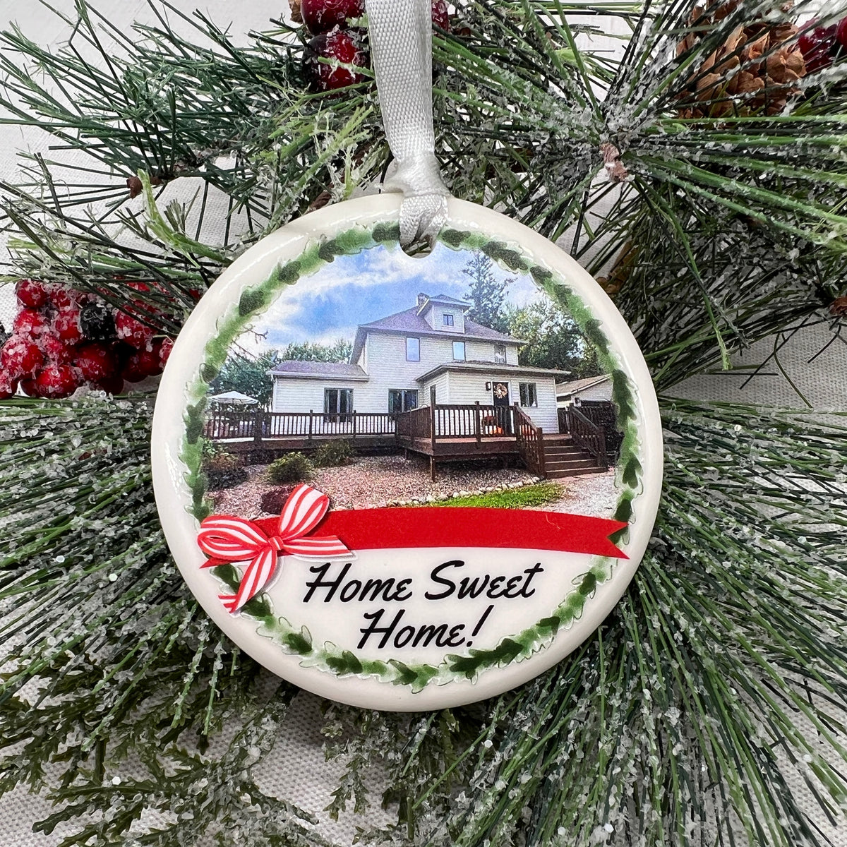 A round ceramic Christmas ornament hanging from a white ribbon, featuring a printed photo of a house, a green wreath border, a red ribbon banner, and the text 'Home Sweet Home!' in black script. The ornament is displayed on a snowy pine tree branch.