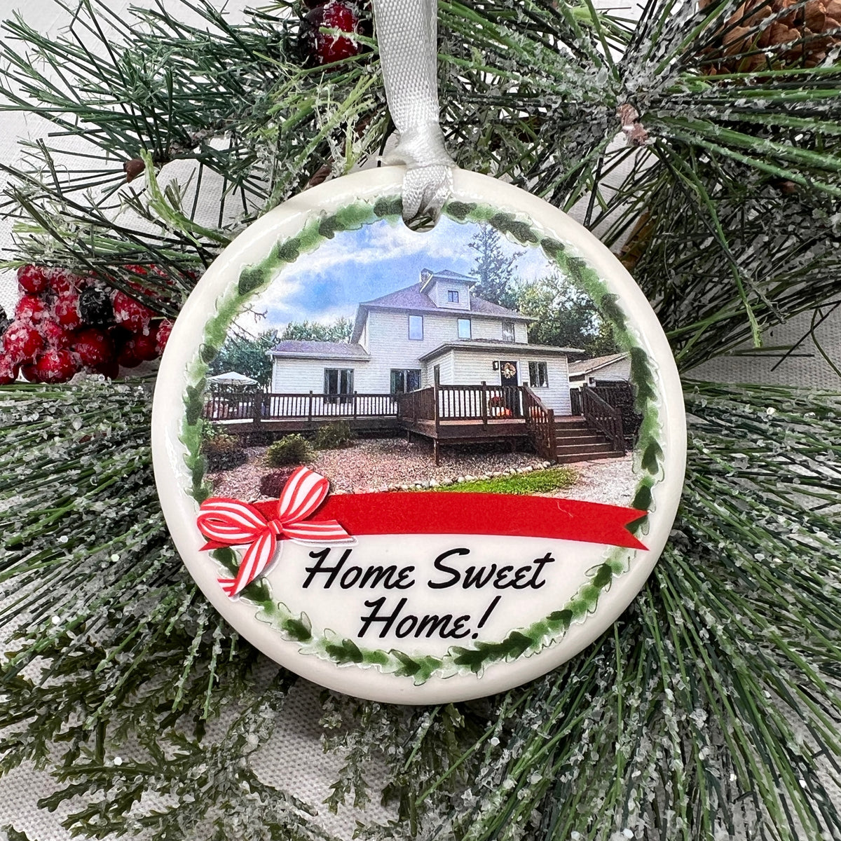 A round ceramic Christmas ornament hanging from a white ribbon, featuring a printed photo of a house, green holly-style border, red ribbon, and the words 'Home Sweet Home!'. The ornament is photographed against frosted evergreen branches with red berries.