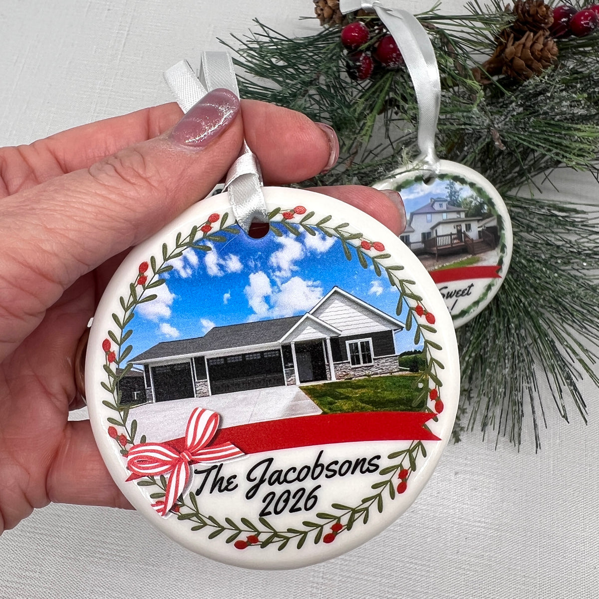 A hand holds a round white ceramic Christmas ornament featuring a custom photo of a house, the text 'The Jacobsons 2026', and a red ribbon bow. Another ornament hangs from a pine branch in the background.