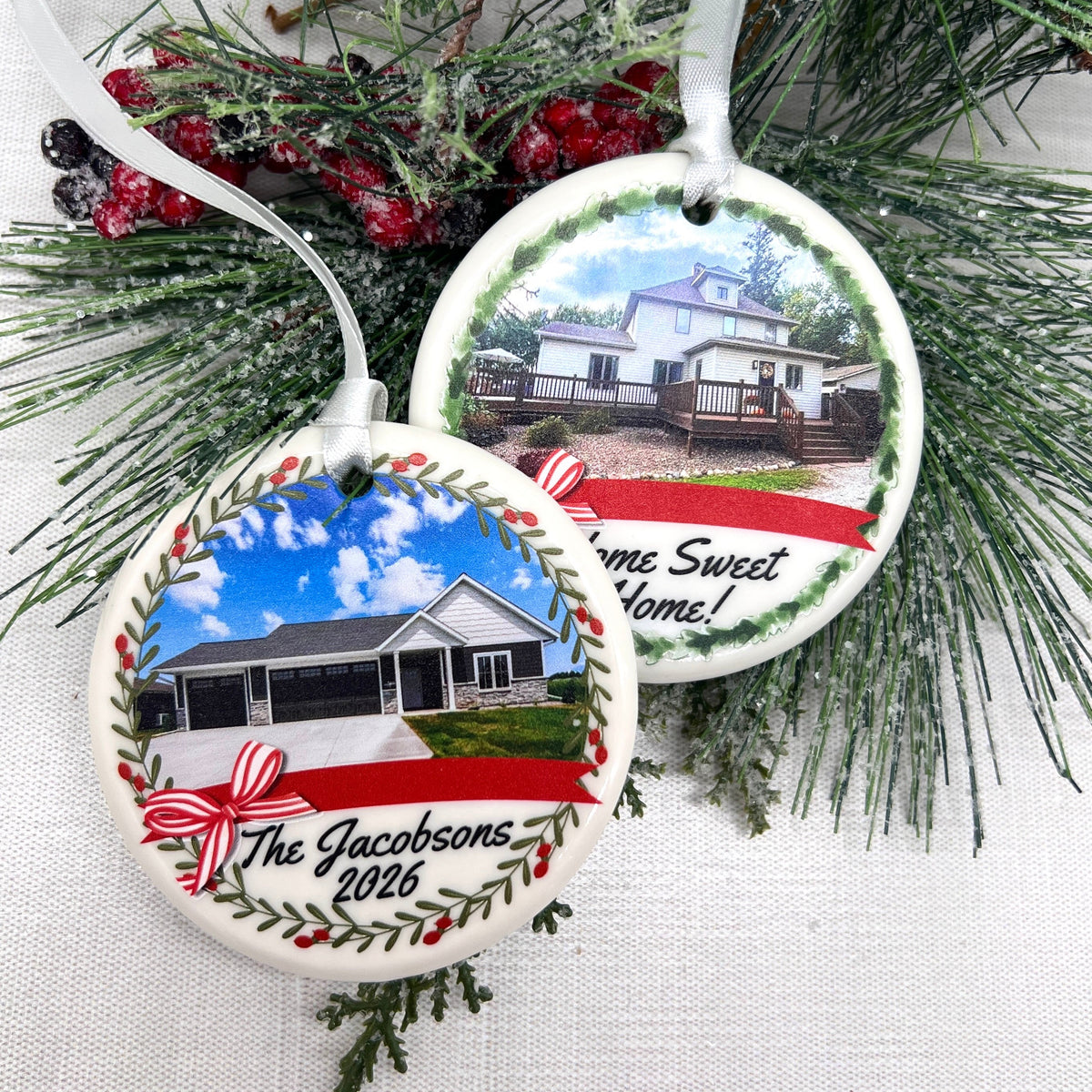 Two personalized round ceramic Christmas ornaments displayed on a white fabric background with green pine branches and red berries. One ornament shows a house photo, a red ribbon, and the text 'The Jacobsons 2026' with a red bow.