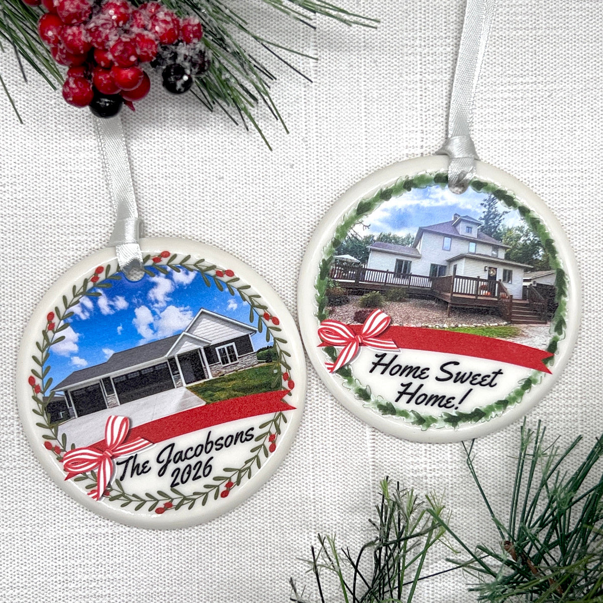 Two round ceramic Christmas ornaments displayed on a white textured fabric background with pine branches and red berries as props. One ornament shows a house photo with the text 'The Jacobsons 2026' and a red ribbon bow, and the other shows a house photo with the text 'Home Sweet Home!' and a red ribbon bow.