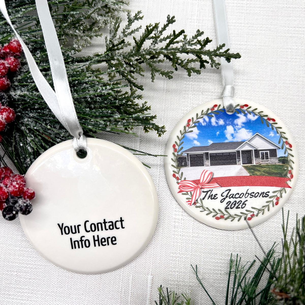 Two round ceramic Christmas ornaments displayed on a white textured fabric with holiday greenery and red berries. One ornament is plain white with black text reading 'Your Contact Info Here' and a white ribbon.