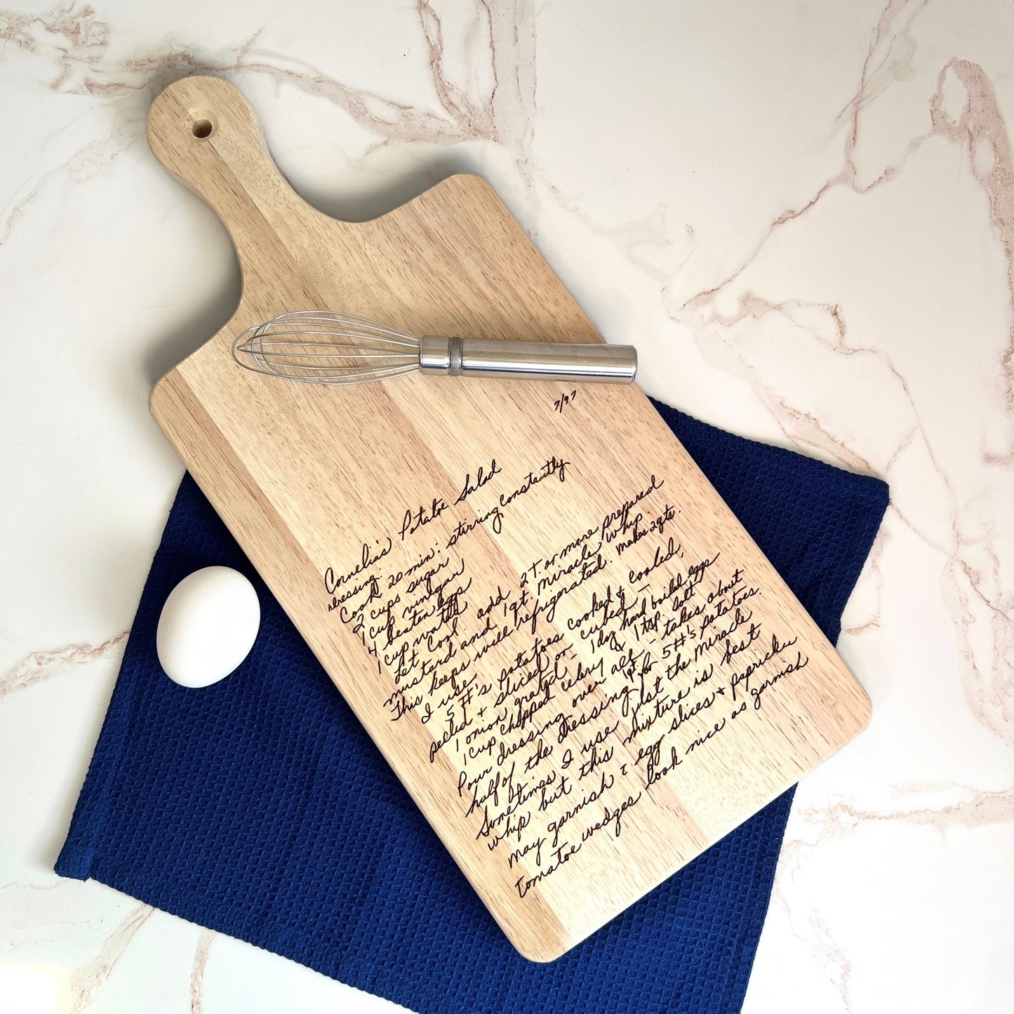 Wooden cutting board with engraved text, whisk, and egg on a blue towel against a marble background
