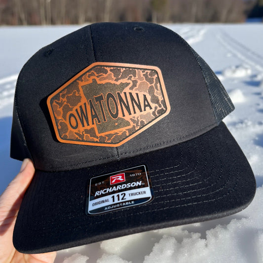 Black Richardson 112 trucker cap  with 'Owatonna' patch with camo background and state of MN, held over a snowy background