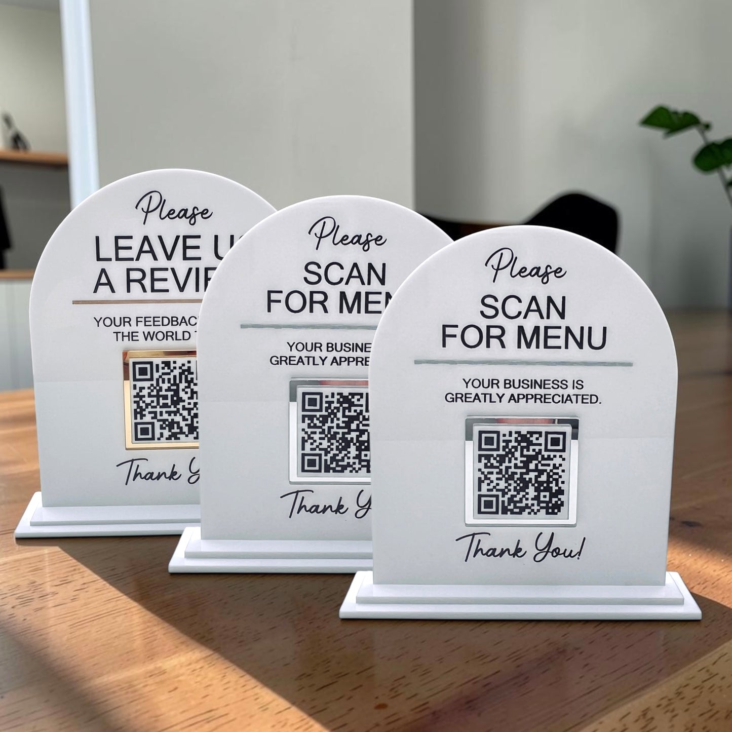 Three white arched tabletop sign holders with QR codes and black text, arranged on a wooden table in a real indoor setting with soft background blur.