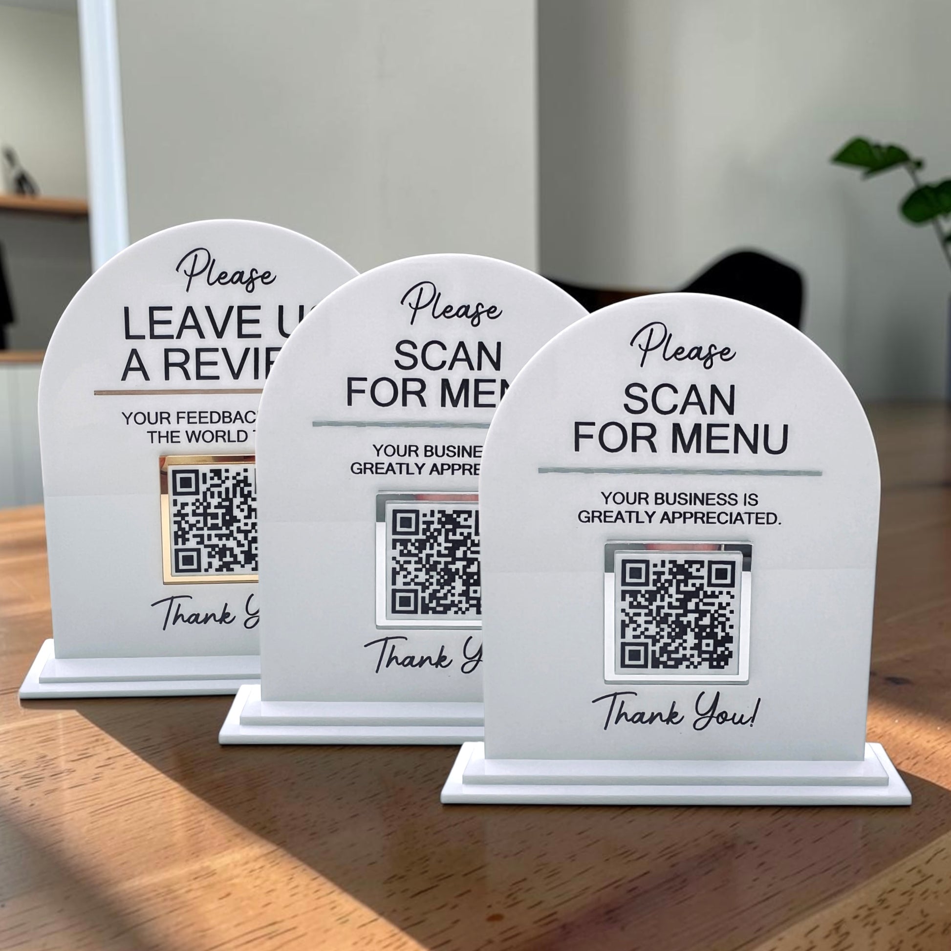 Three white arched tabletop sign holders with QR codes and black text, arranged on a wooden table in a real indoor setting with soft background blur.