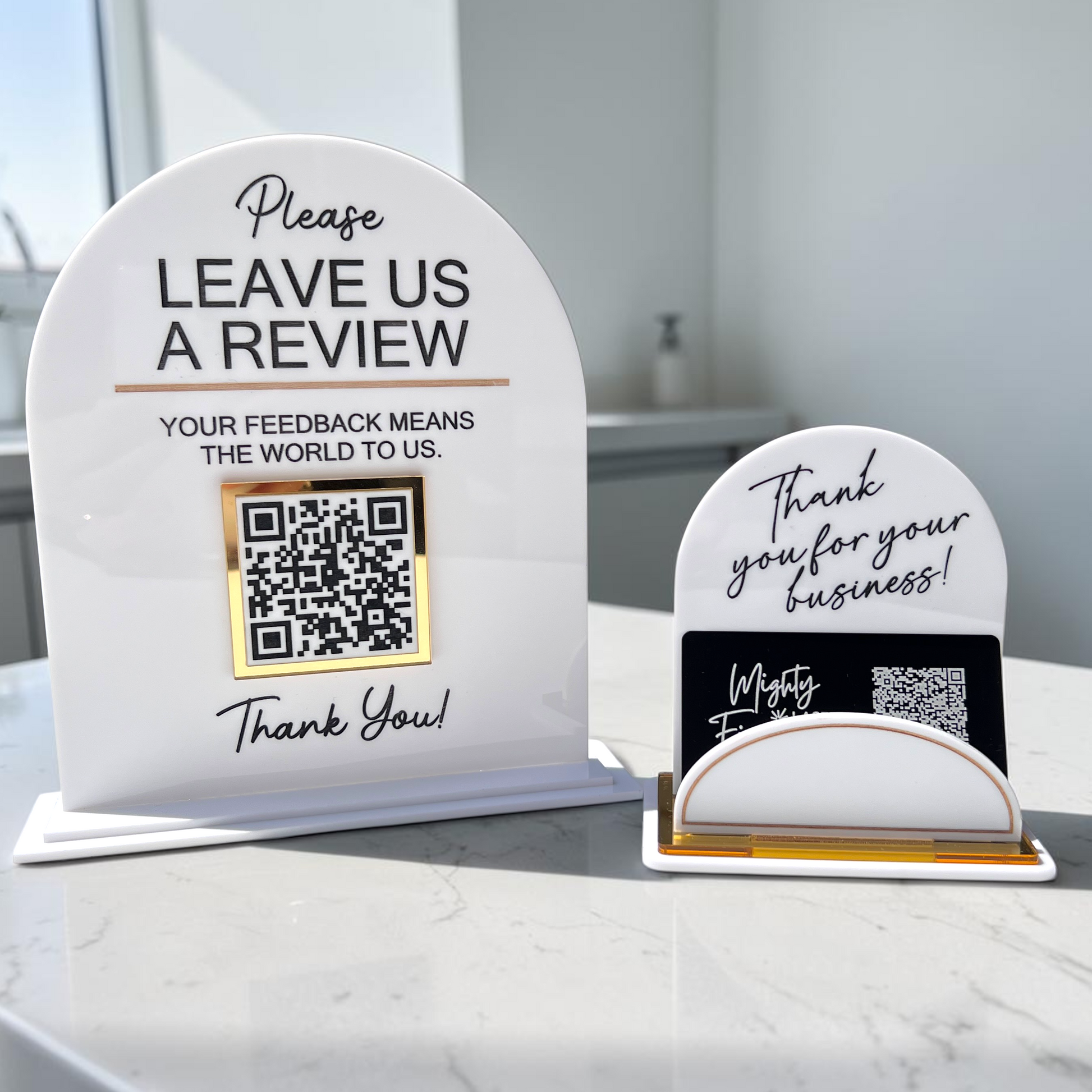 Two white acrylic tabletop review signs on a marble countertop in a bright interior. The larger sign says 'Please LEAVE US A REVIEW' with a QR code, and the smaller sign says 'Thank you for your business!' with a business card holder.