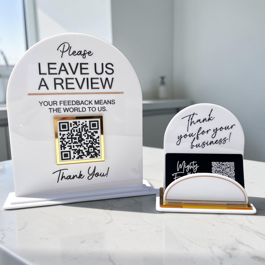 Two white acrylic tabletop review signs on a marble countertop in a bright interior. The larger sign says 'Please LEAVE US A REVIEW' with a QR code, and the smaller sign says 'Thank you for your business!' with a business card holder.