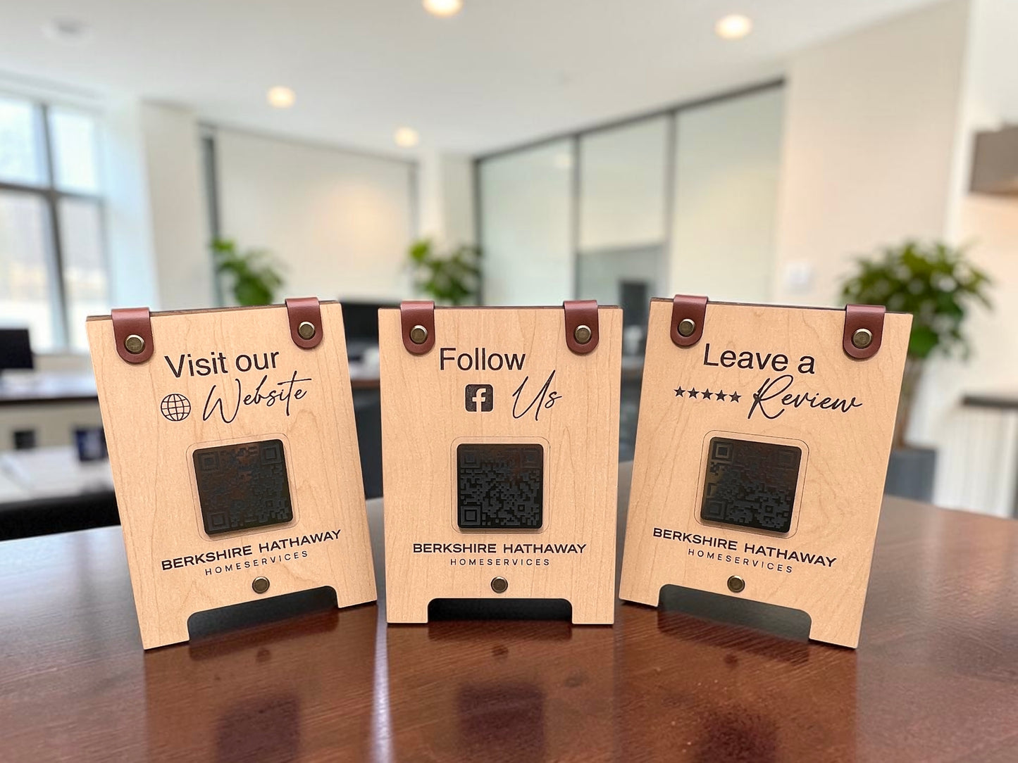 Three wooden QR code review signs with brown leather hinges and engraved text reading 'Visit our Website', 'Follow Us', and 'Leave a Review', displayed on a wooden table in a modern office setting.