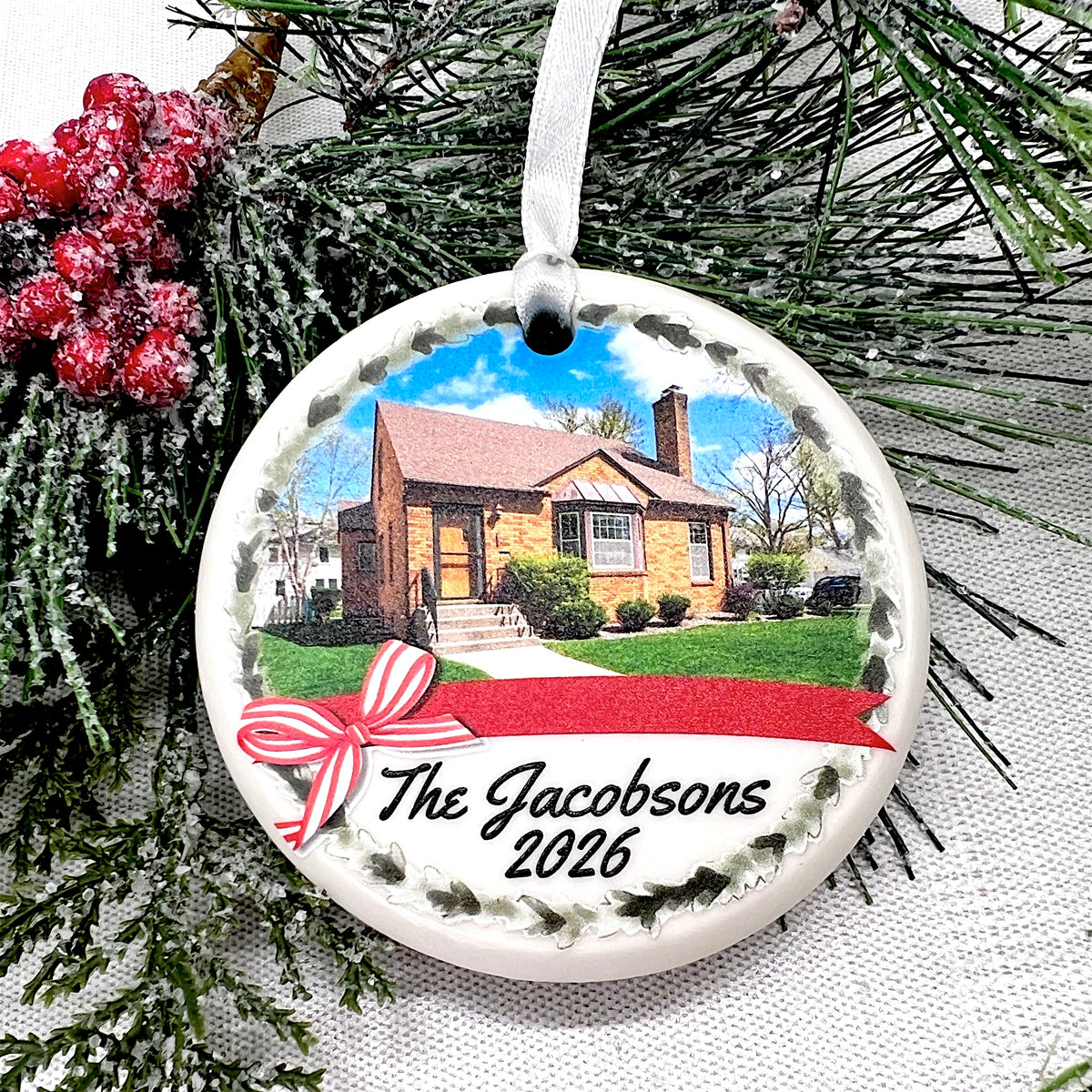 A personalized round ceramic Christmas ornament featuring a printed photo of a house, a red ribbon bow, and the text 'The Jacobsons 2026', hanging against evergreen branches and red berries.