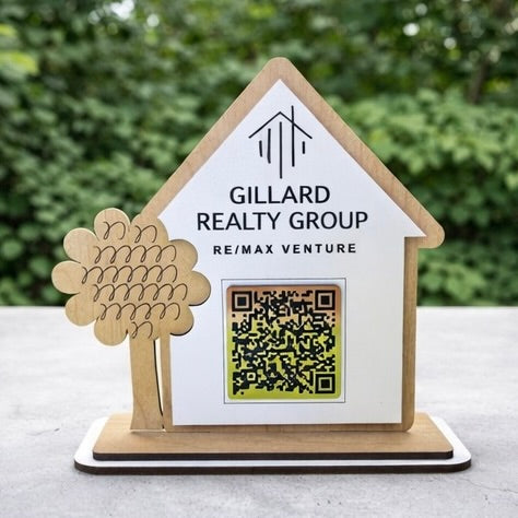 Wooden house-shaped sign with 'Gillard Realty Group' branding and a QR code on a blurred green background.
