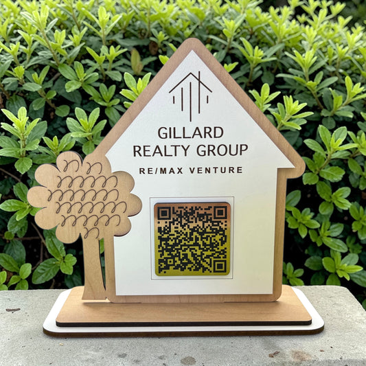 Wooden house-shaped sign with 'Gillard Realty Group' branding and a QR code, against a green foliage background.