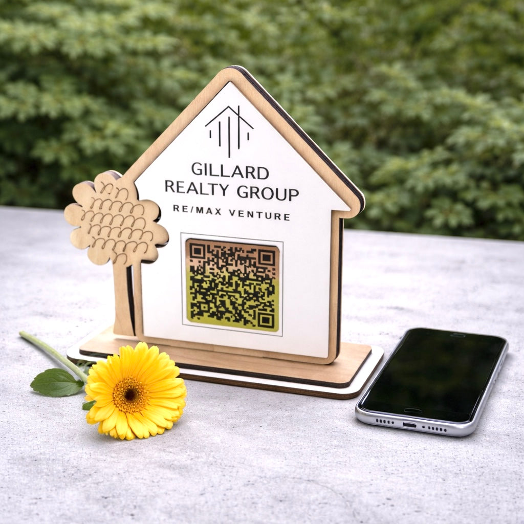 Gillard Realty Group sign with QR code on a stand next to a smartphone and yellow flower.