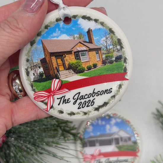 A round ceramic ornament with a picture of a brick home and The Jacobsons 2026 on the front. Held with a hand with another ornament and decorative green pines in the background.