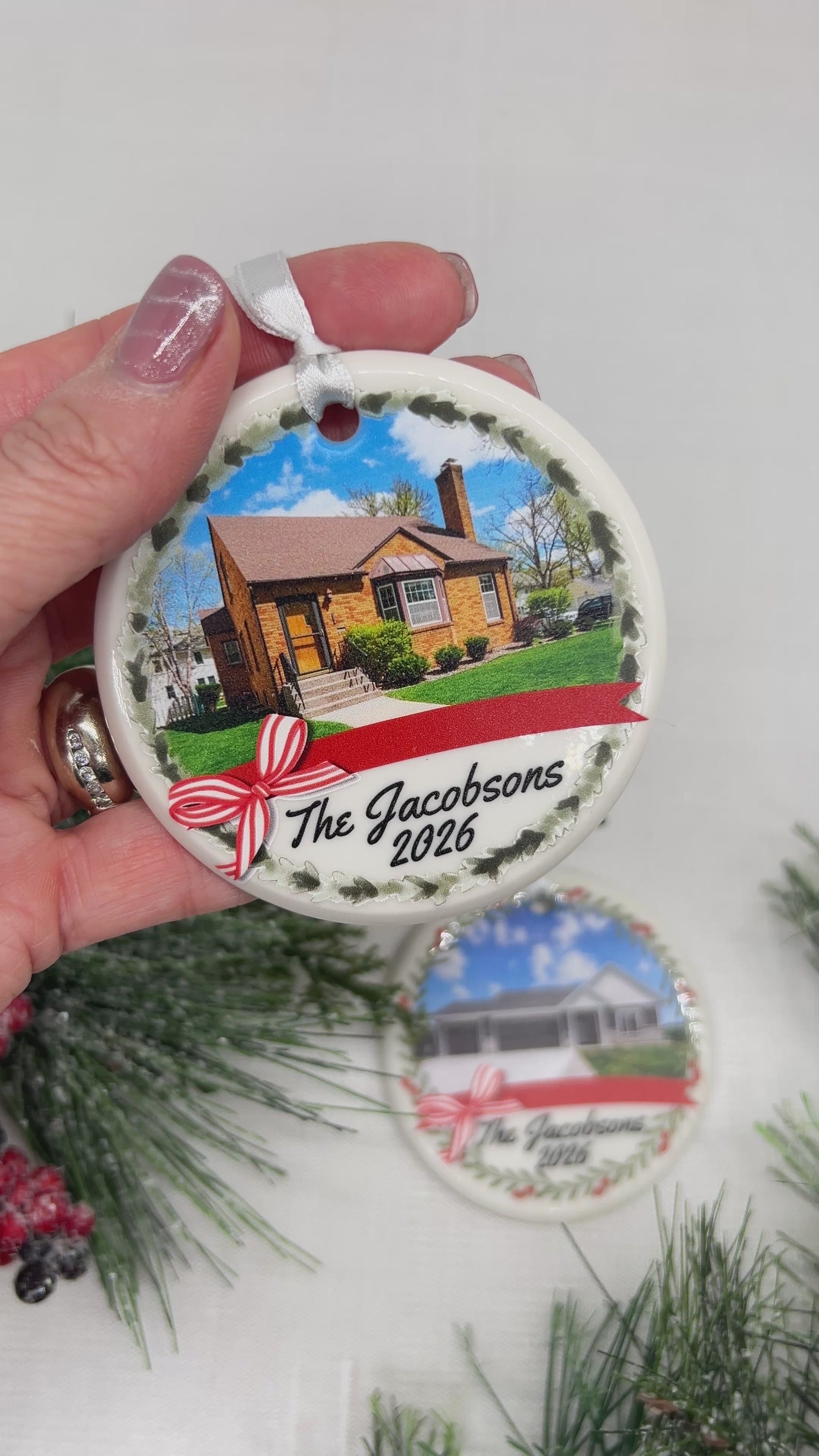 A round ceramic ornament with a picture of a brick home and The Jacobsons 2026 on the front. Held with a hand with another ornament and decorative green pines in the background.
